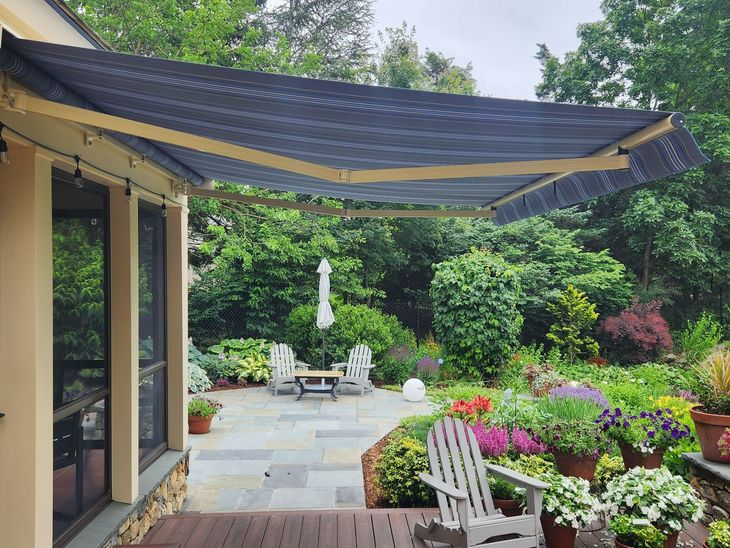 Retractable blue and beige awning extends over a patio with a garden. MasterCraft Roofing, Siding & Windows | Serving Eastern MA, NH & Selected Areas of VT