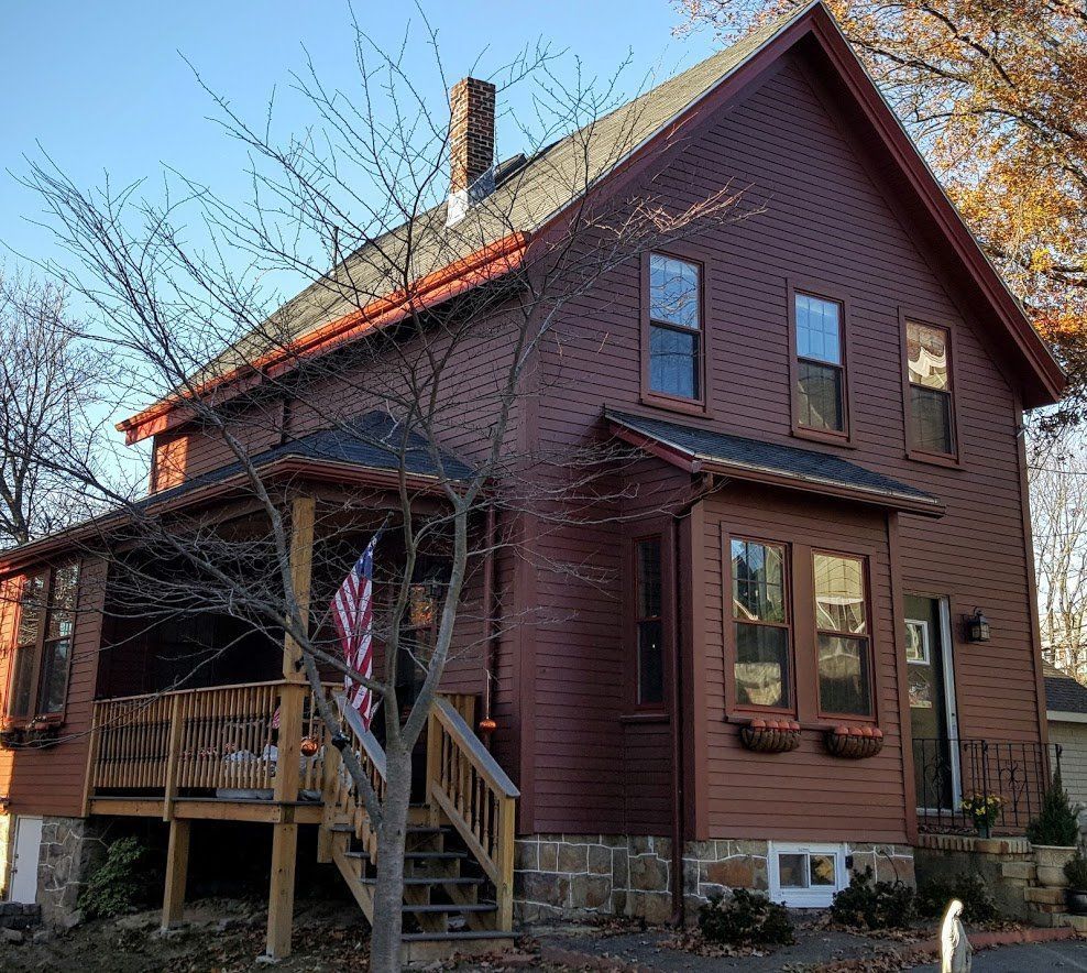 Brown two-story house with porch, steep roof, and bare trees in front