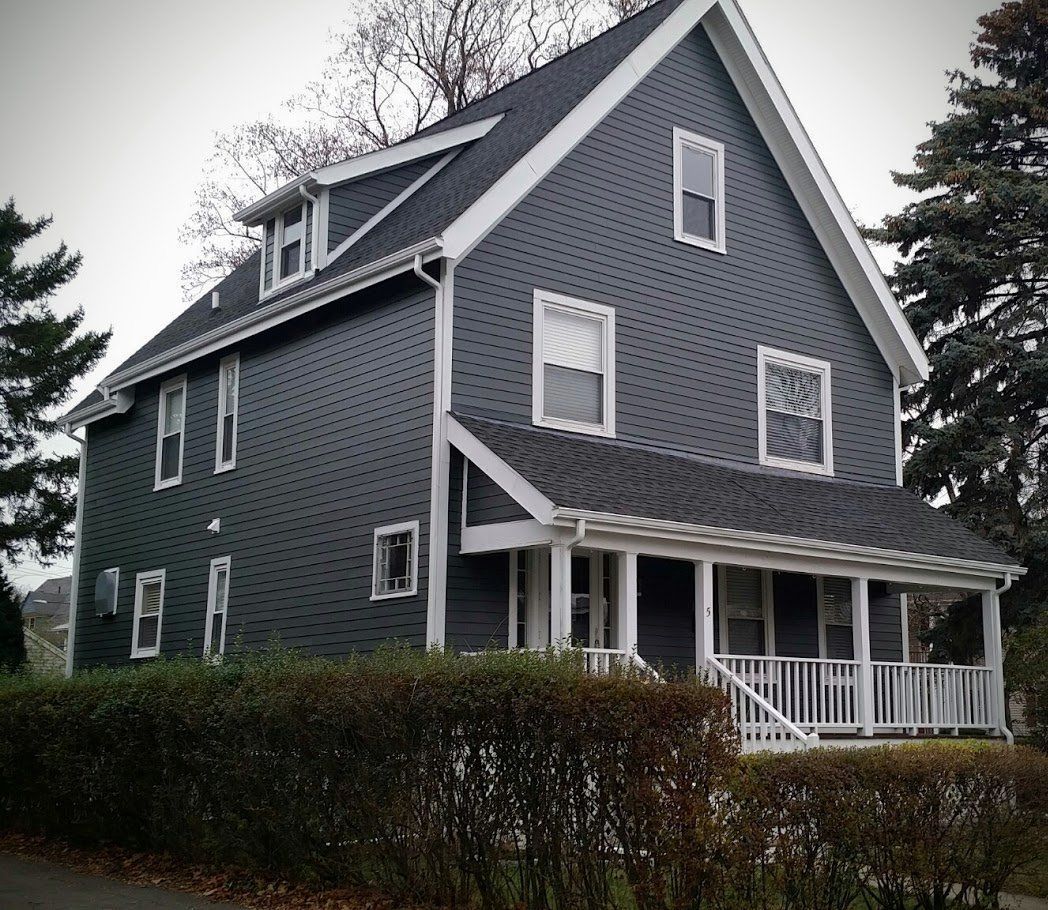 Gray two-story house with white trim, steep roof, and front porch behind a hedge