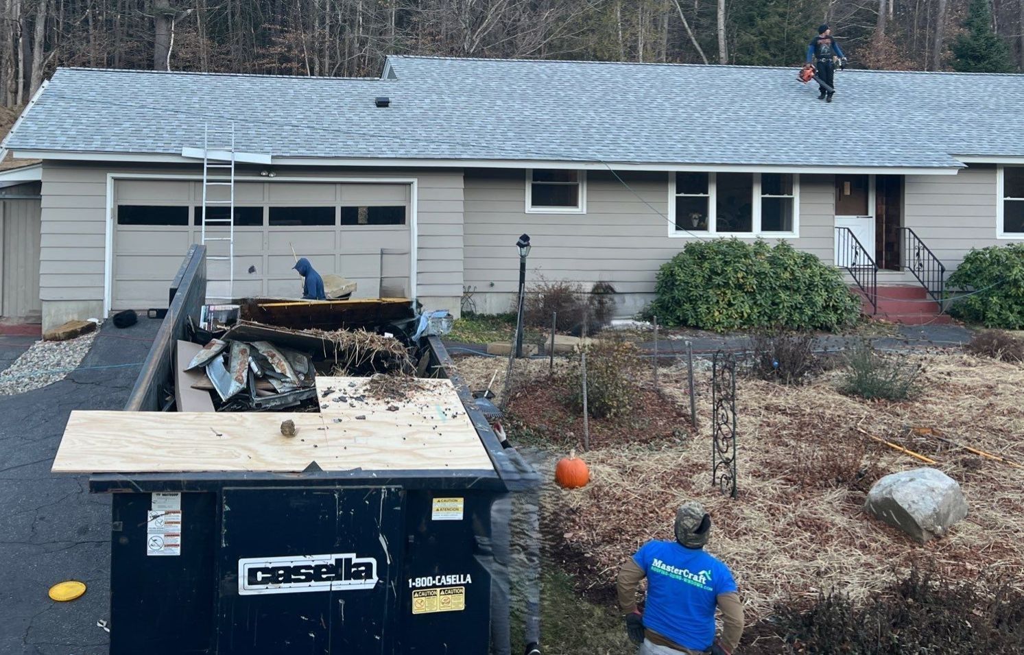 Roofers working on a house with a dumpster in the driveway. One person is on the roof, others near the dumpster.
