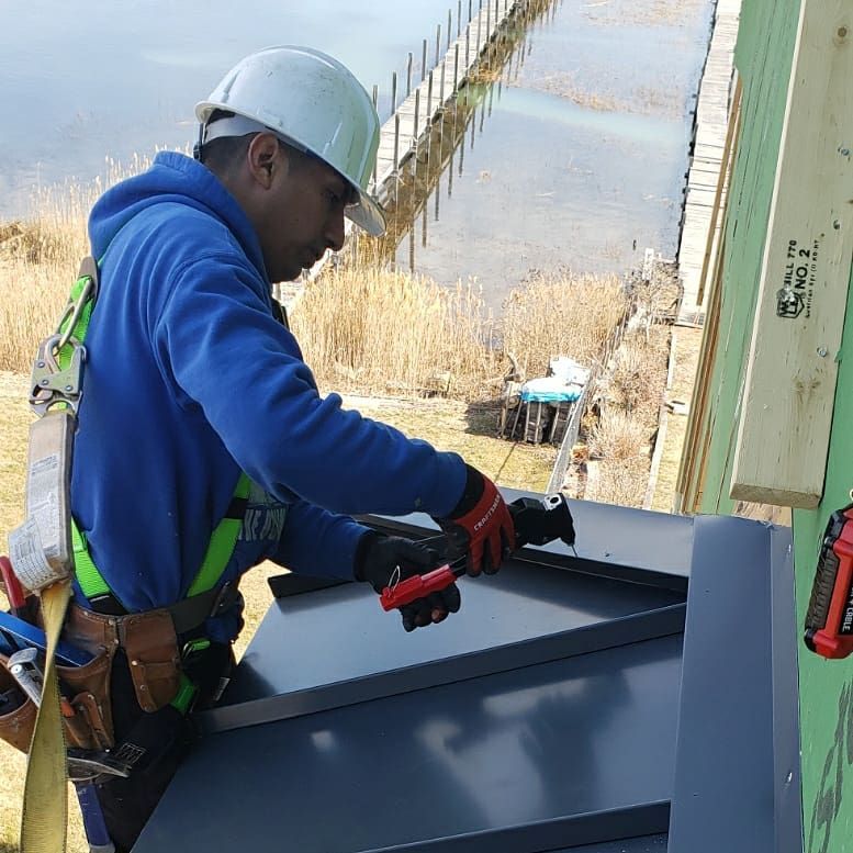 Roofer in safety gear installing metal roofing sheets outdoors near water. MasterCraft Roofing, Siding & Windows | Serving Eastern MA, NH & Selected Areas of VT