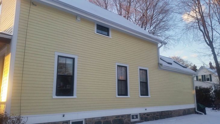 Yellow house with black window frames, snow on the roof, winter setting. MasterCraft Roofing, Siding & Windows | Serving Eastern MA, NH & Selected Areas of VT