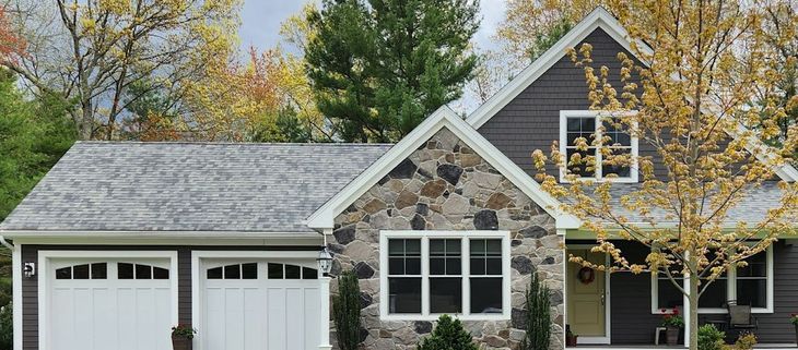 Stone and gray house with white garage doors and a yellow tree. MasterCraft Roofing, Siding & Windows | Serving Eastern MA, NH & Selected Areas of VT