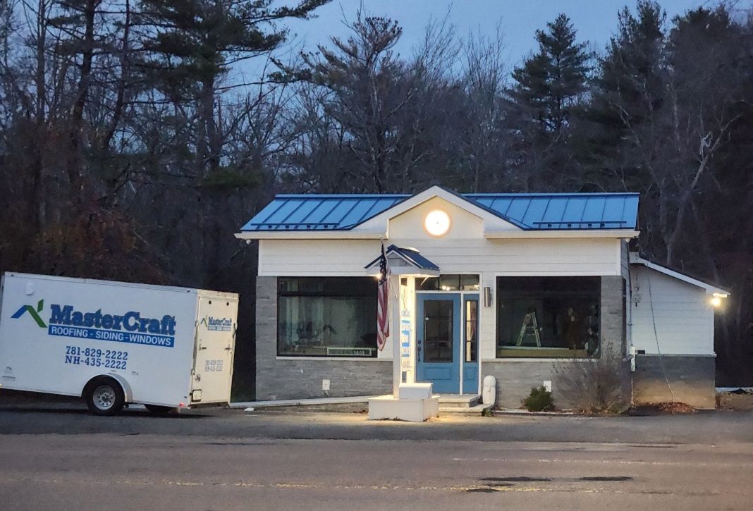 Small white roadside building with blue roof at dusk, beside a utility trailer and wooded background.