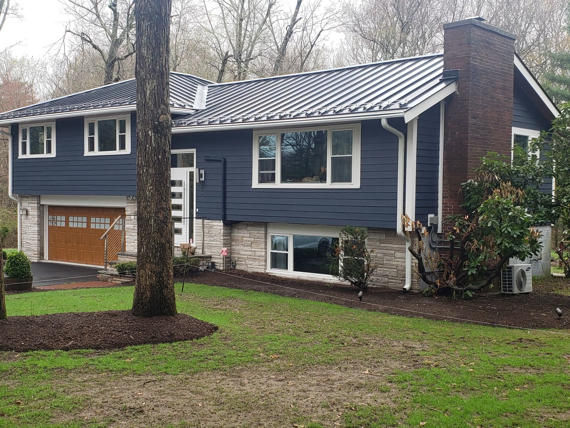 Blue house with brown garage door, brick chimney, and metal roof. Landscaped front yard with a tree. Siding | MasterCraft Roofing, Siding & Windows | Serving Eastern MA, NH & Selected Areas of VT
