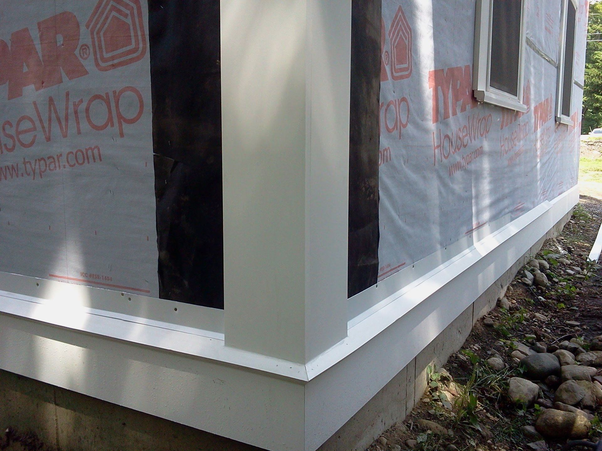 Corner of a house with gray siding, white trim, and exposed house wrap over a rocky foundation.