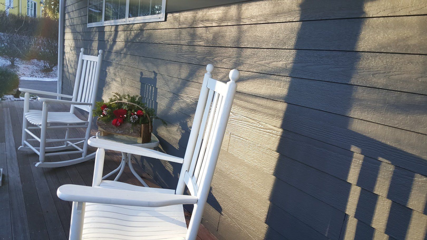 White rocking chairs on a sunlit porch beside a small table with red flowers and a brick wall.
