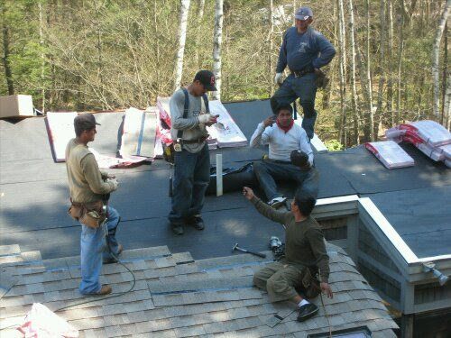 Roofers on a rooftop, working and taking a break. Bags of shingles are nearby. Sunlight and trees in the background.