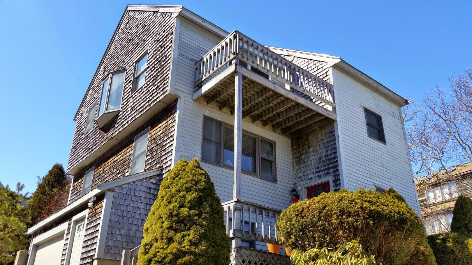 Three-story house with white siding, stone facade, and a balcony, framed by trimmed shrubs under a blue sky