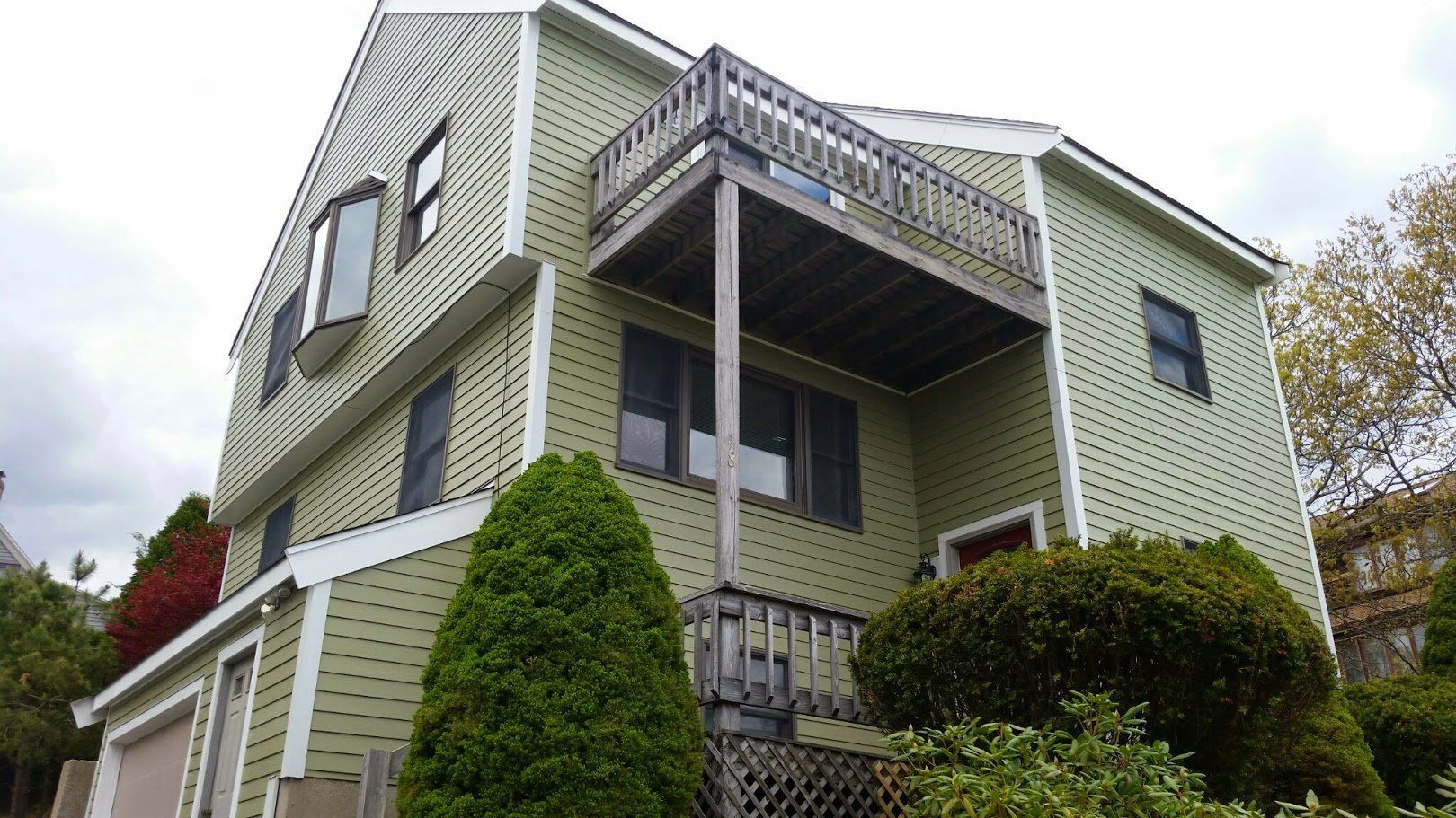 Three-story house with green siding, balcony, and surrounding shrubs viewed from below.