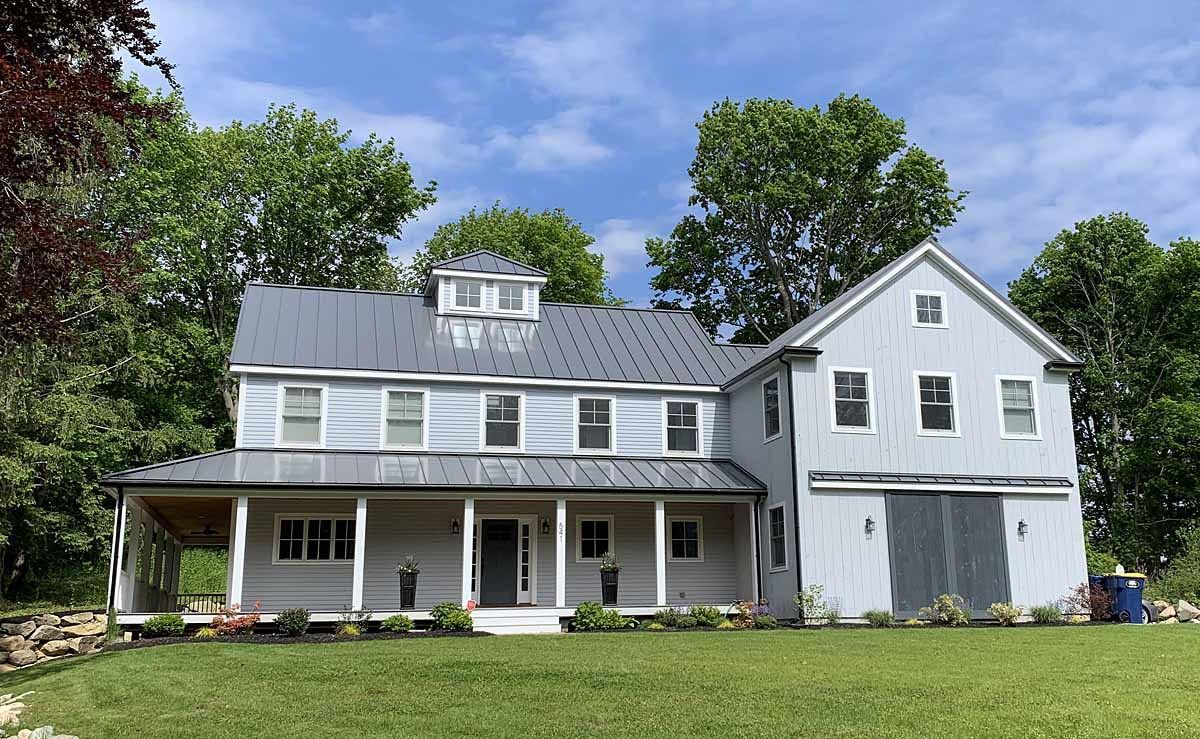 White two-story suburban house with porch, gray roof, and attached garage surrounded by trees and lawn
