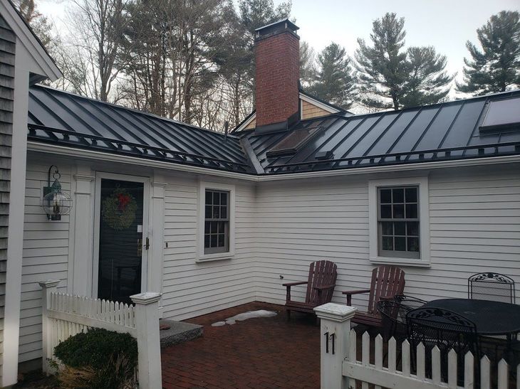 White house with black metal roof, brick chimney, small front patio, and red Adirondack chairs. MasterCraft Roofing, Siding & Windows | Serving Eastern MA, NH & Selected Areas of VT
