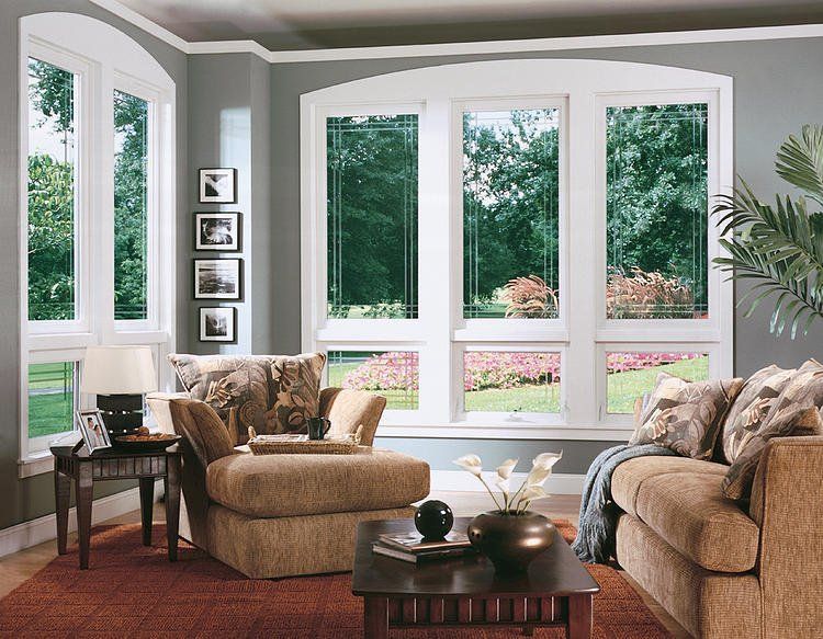 Bright living room with large windows, beige chairs, dark coffee table, and a view of trees and flowers outside