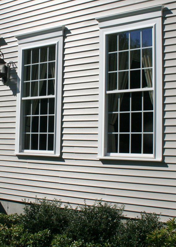 Two white-framed windows on a beige house exterior with horizontal siding and shrubs below.