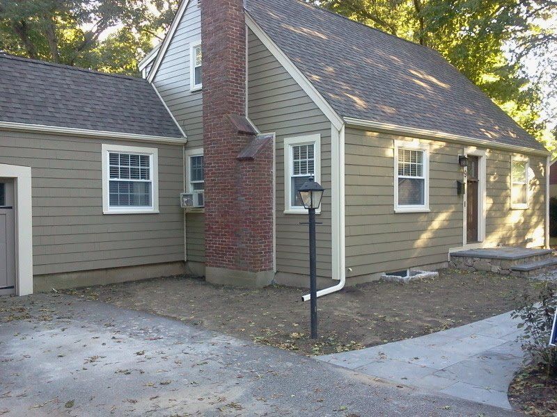 Small tan house with red brick chimney, white trim, and a front walkway in a wooded yard.