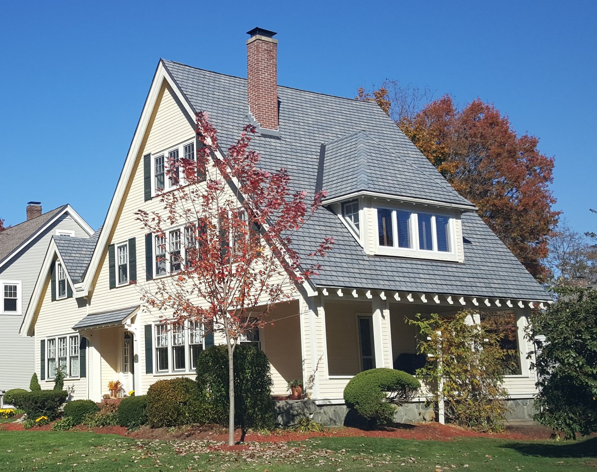 Yellow house with gray roof, porch, and chimney against a blue sky. Roofing | MasterCraft Roofing, Siding & Windows | Serving Eastern MA, NH & Selected Areas of VT