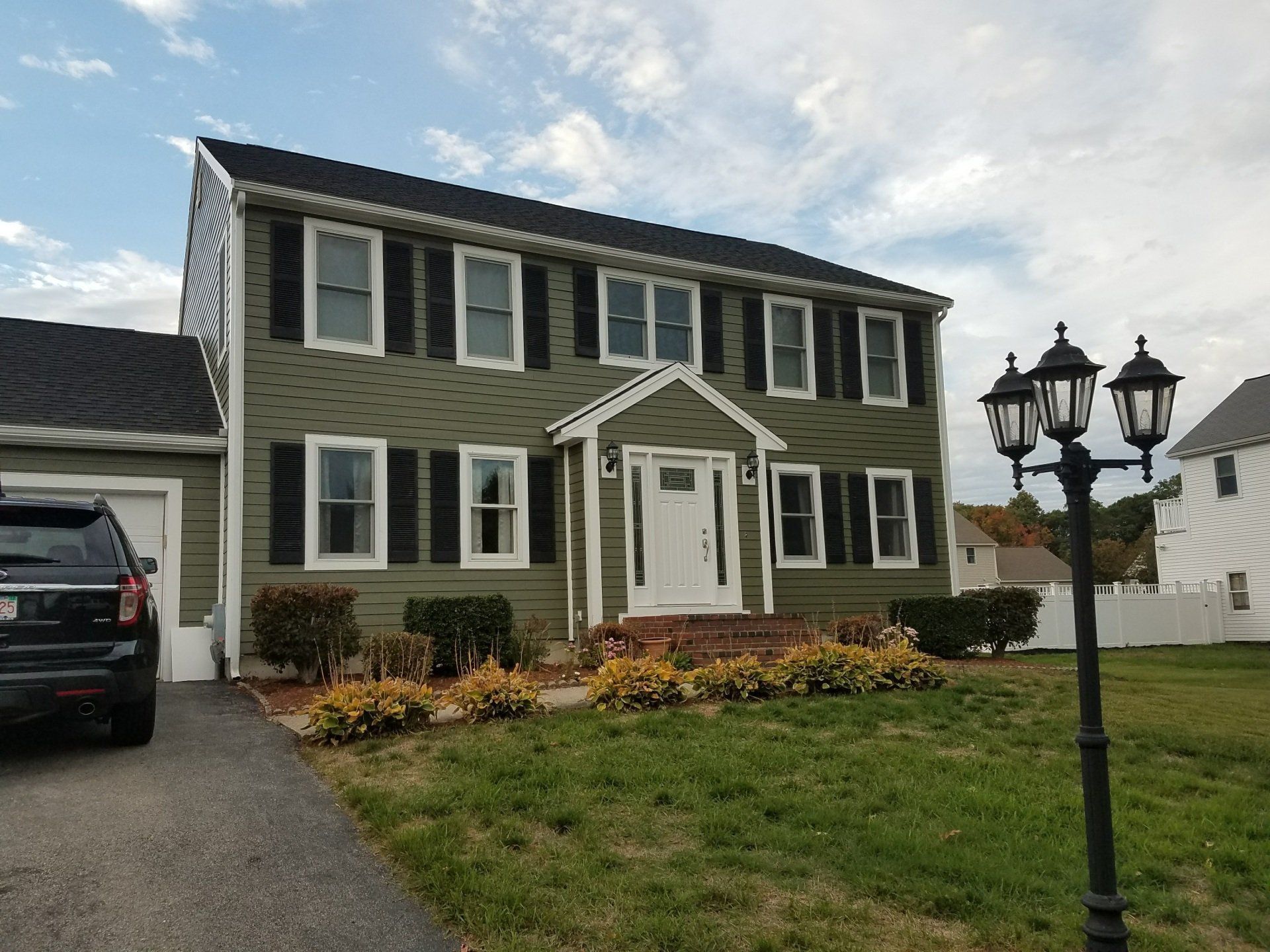 Suburban two-story house with green siding, white trim, a driveway, lawn, and black lamp post in front
