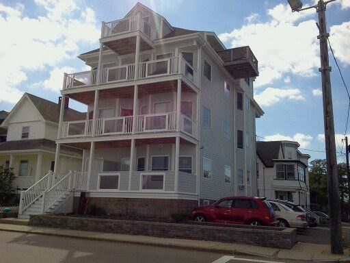 Three-story Victorian-style house with porches and a red car parked beside it under a partly cloudy sky