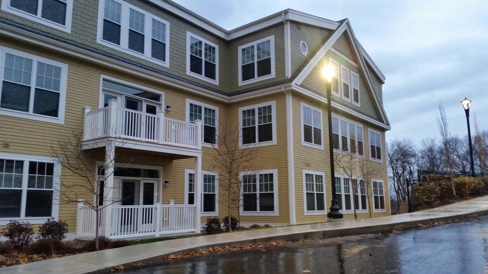 Modern apartment building with tan siding, white balconies, and lit streetlamp on a wet street