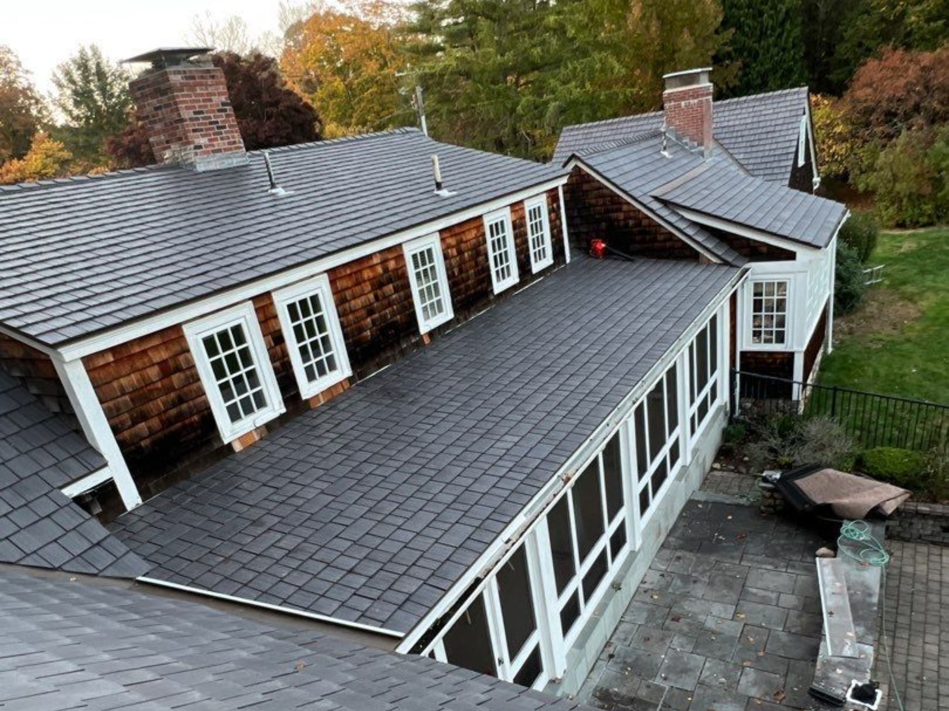 Dark slate-tiled roof on a long house with white-framed windows, brick chimney, and surrounding greenery. Roofing | MasterCraft Roofing, Siding & Windows | Serving Eastern MA, NH & Selected Areas of VT