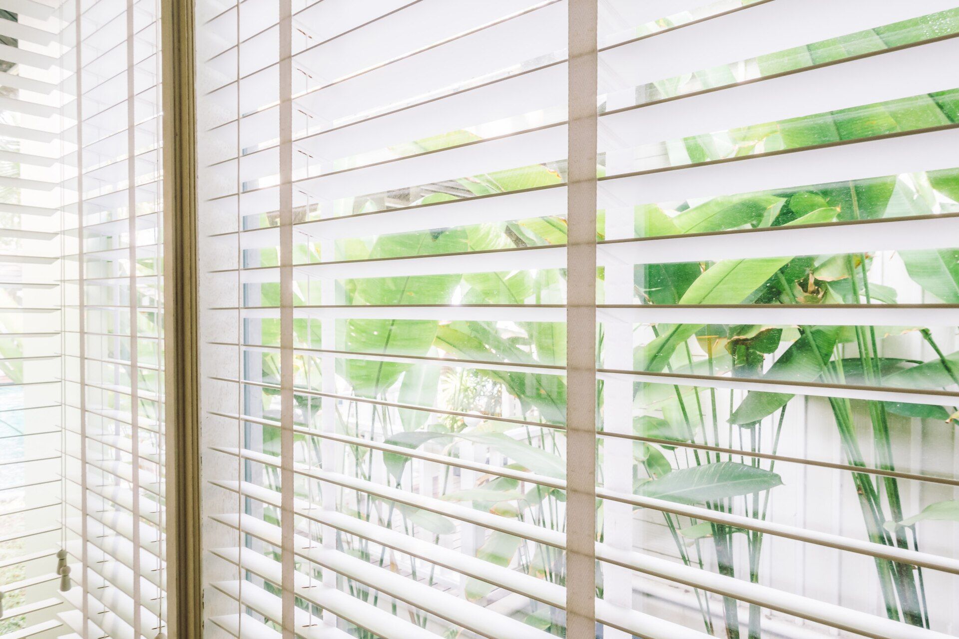Wooden blinds, partially open, provide a view of lush greenery outside a window. Shades & Awnings | MasterCraft Roofing, Siding & Windows | Serving Eastern MA, NH & Selected Areas of VT