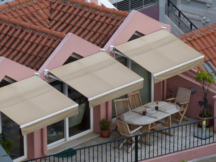 A terrace with tan awnings, a table, and chairs on a pink building with red tile roofing. Shades & Awnings | MasterCraft Roofing, Siding & Windows | Serving Eastern MA, NH & Selected Areas of VT