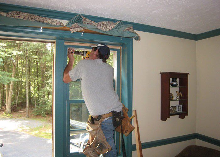 Man installing window frame, indoors. He's using a tool, with a forest view outside. Room has teal trim and a small cabinet. Windows & Doors | MasterCraft Roofing, Siding & Windows | Serving Eastern MA, NH & Selected Areas of VT