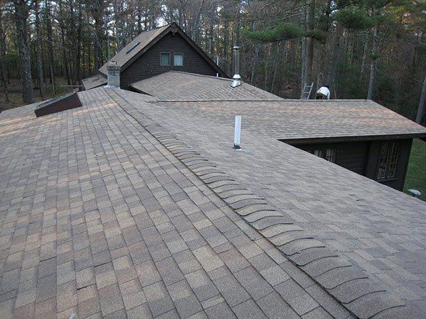 Brown shingled roof of a house with multiple sections and chimneys, set in a wooded area. Roof Repairs | MasterCraft Roofing, Siding & Windows | Serving Eastern MA, NH & Selected Areas of VT