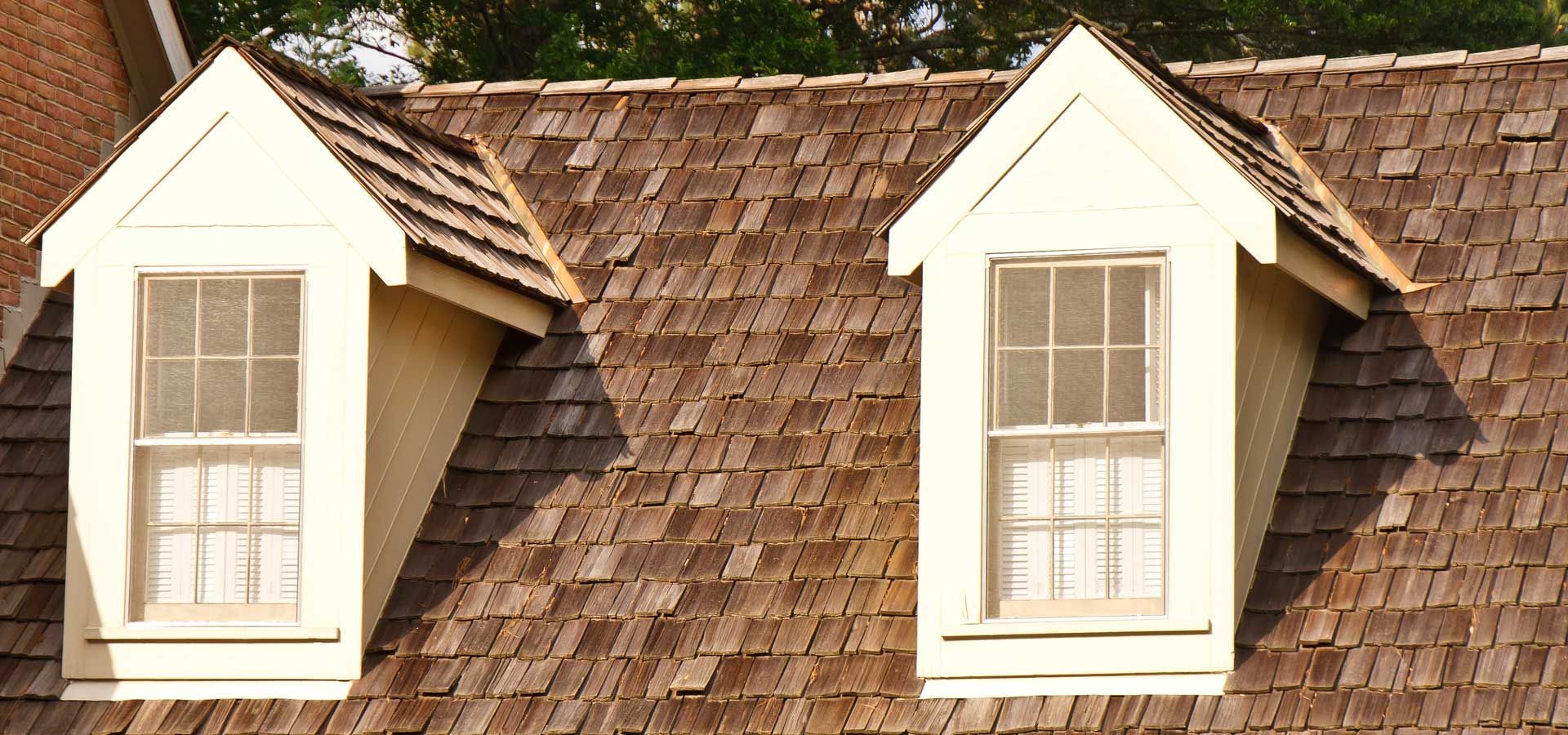 Two dormer windows on a cedar shake roof, beige frames, tan siding, against a green background. Roofing | MasterCraft Roofing, Siding & Windows | Serving Eastern MA, NH & Selected Areas of VT