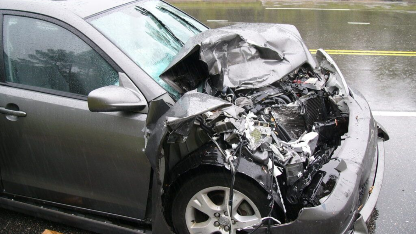 Damaged gray car front-end, crumpled from a collision on a wet road.