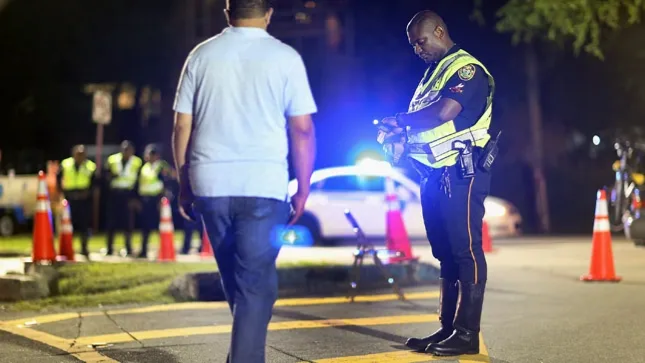 A police officer checks a man during a nighttime DUI checkpoint; other officers and a patrol car are in the background.