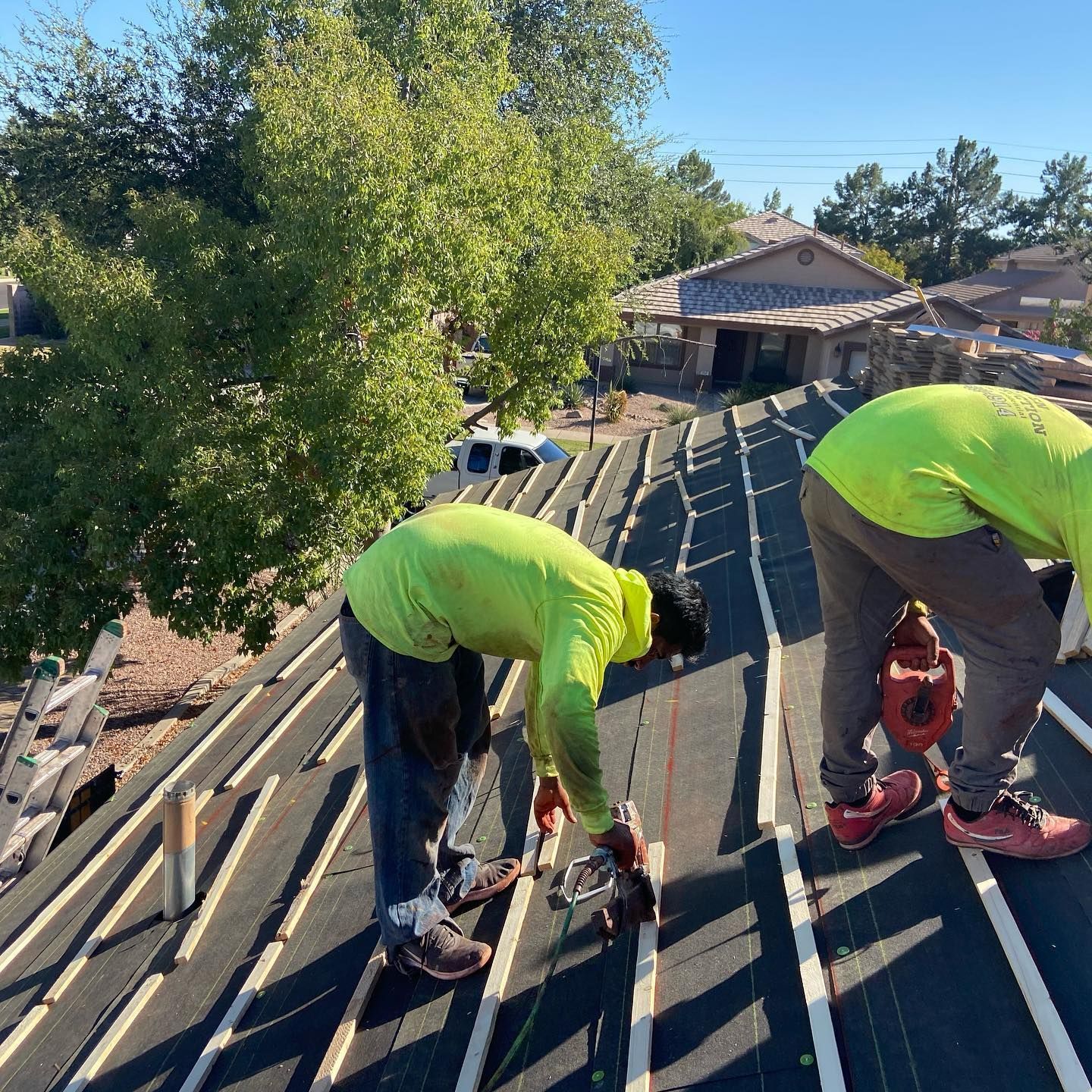 Two roofers in neon shirts work on a rooftop with a house and trees in the background.