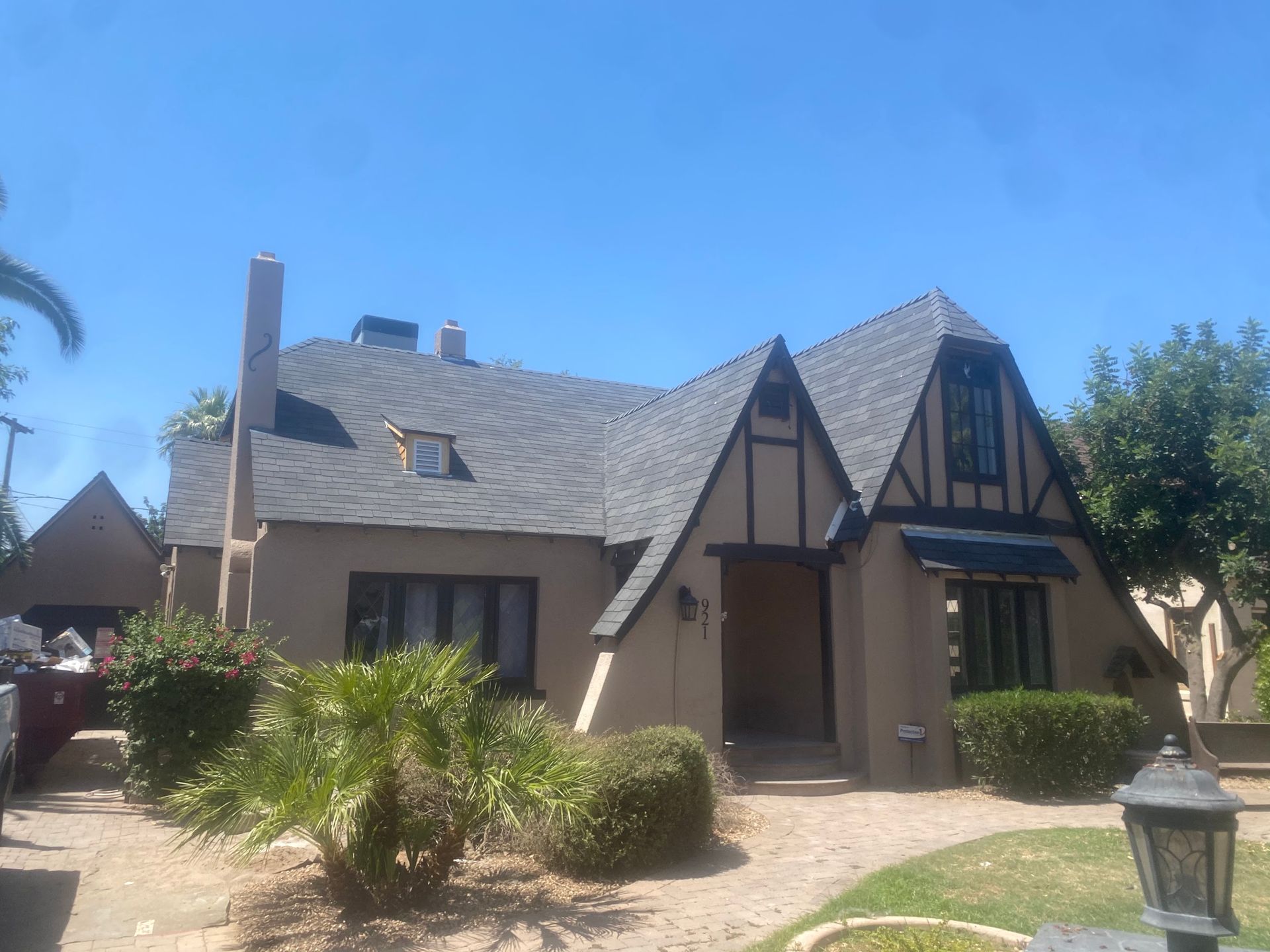 Tan Tudor-style house with dark trim and a gray roof under a blue sky, front yard with landscaping.