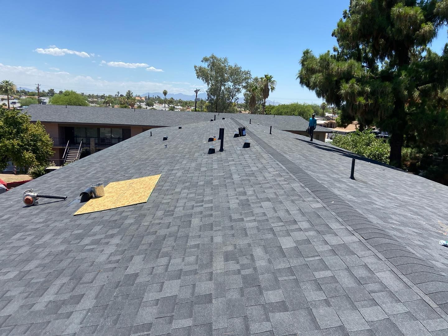 View of a partially completed dark shingle roof with vents and a worker, under a blue sky.