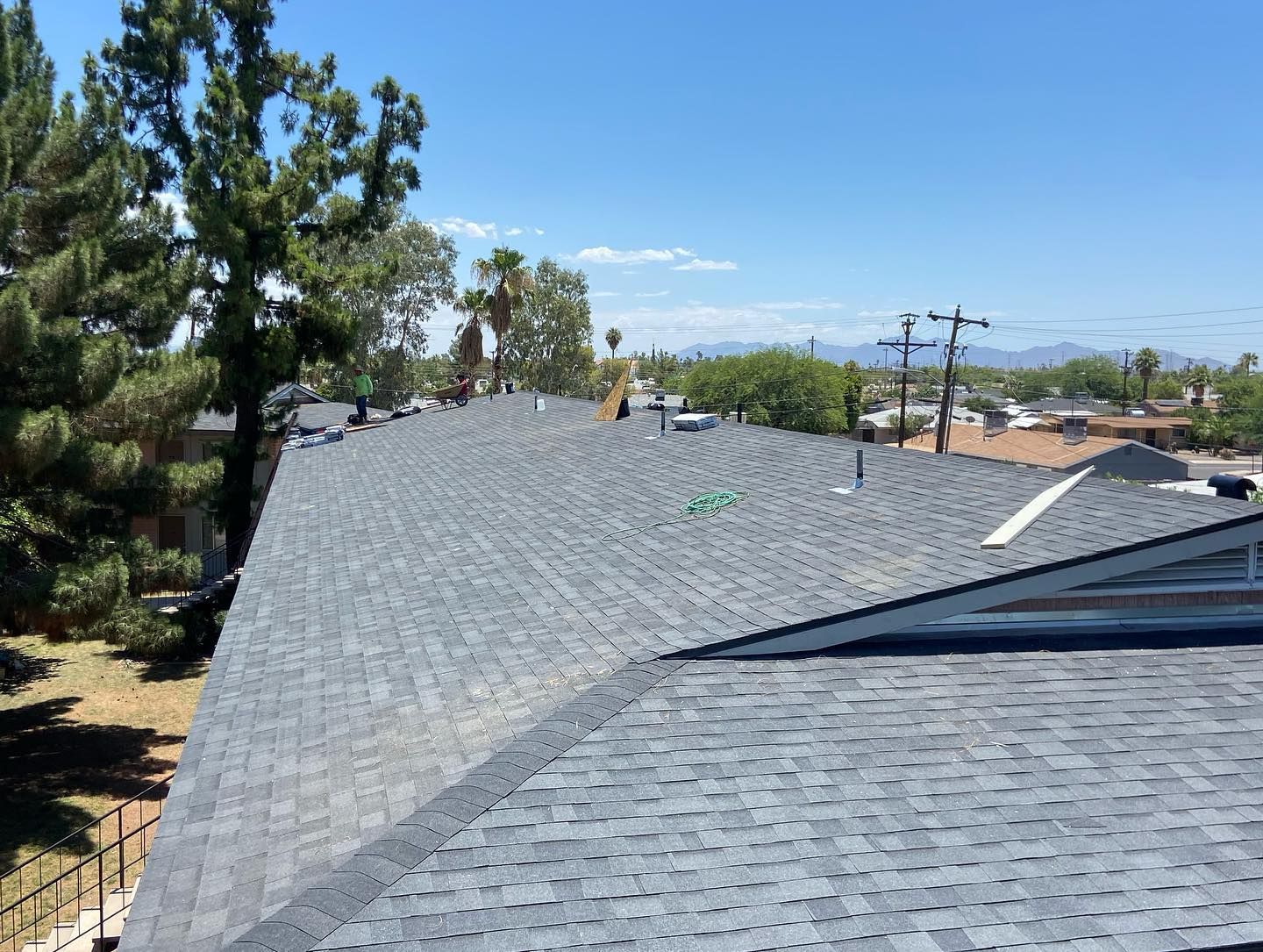 Newly shingled, gray roof on a sunny day. Trees and houses in the background. Blue sky.