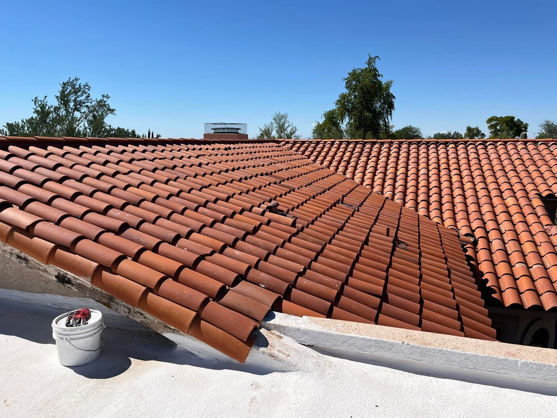 Red tile roof being repaired with a bucket of tools on a white surface, blue sky.