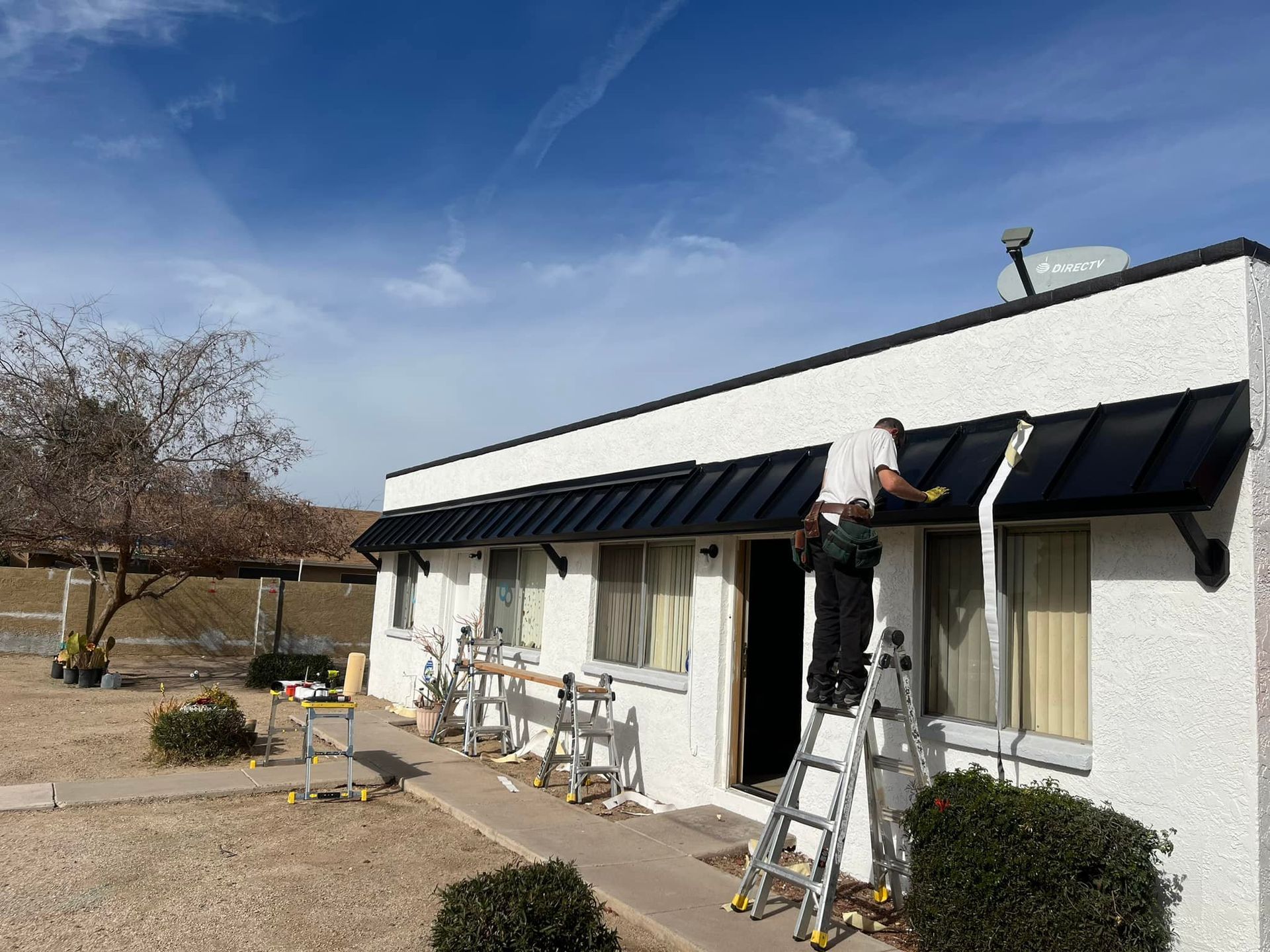 A person on a ladder installs black awning on a white building under a blue sky.