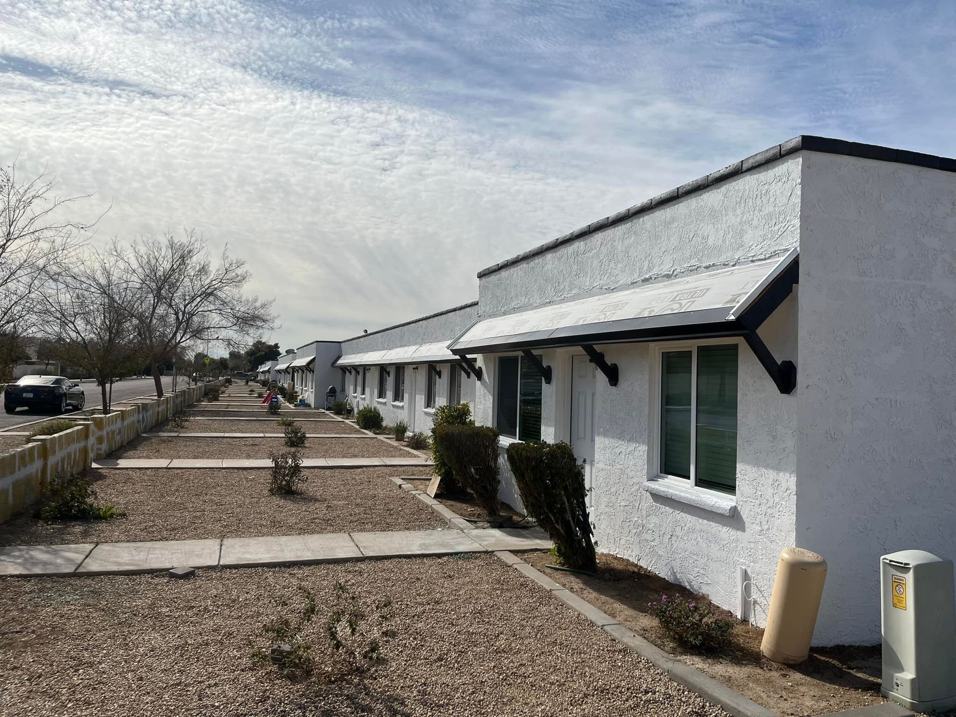 Row of white, stucco-style buildings with awnings, sidewalk with bushes and trees, sunny day.