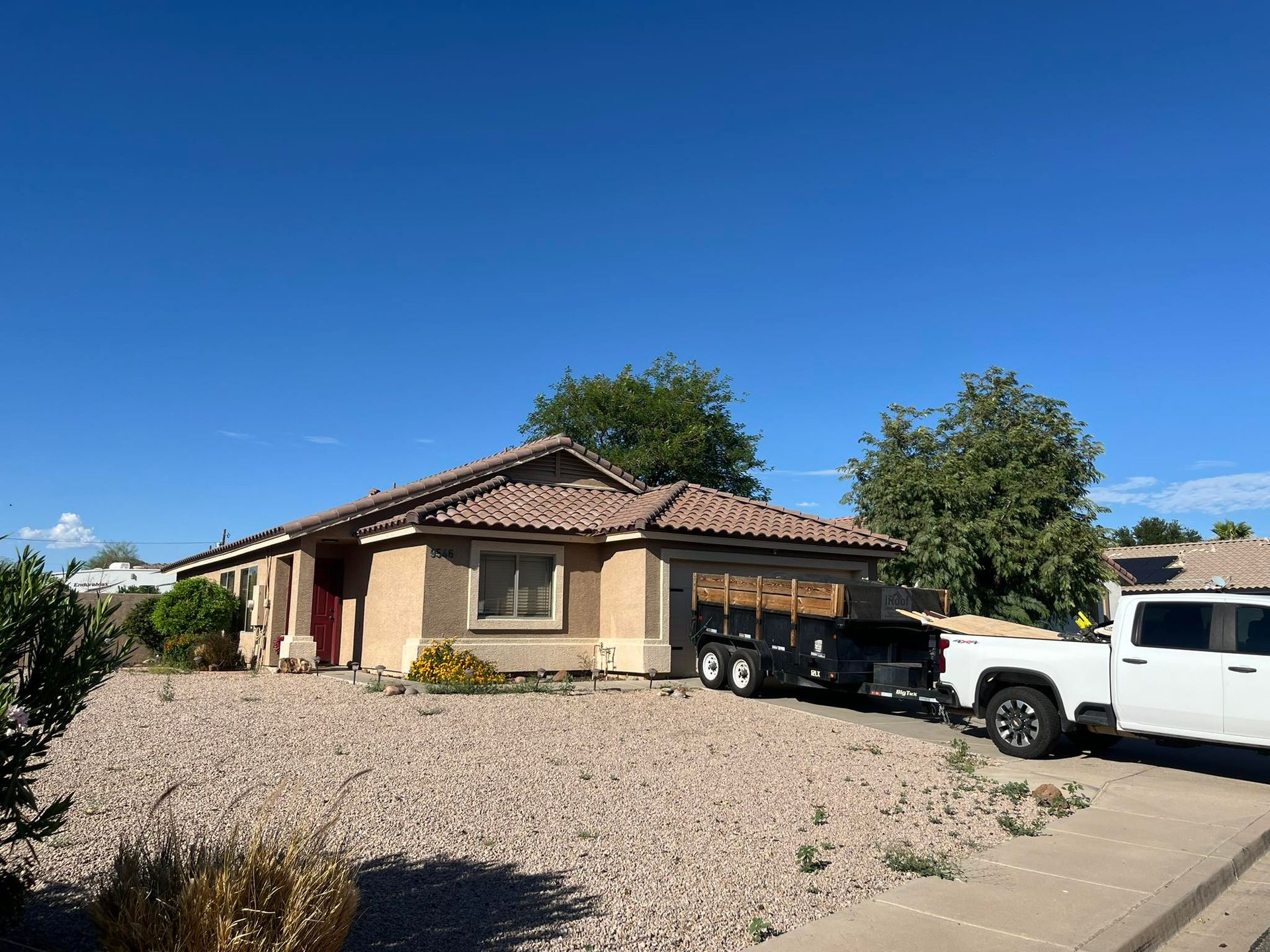 Tan house with a white pickup truck pulling a trailer on a gravel yard under a blue sky.