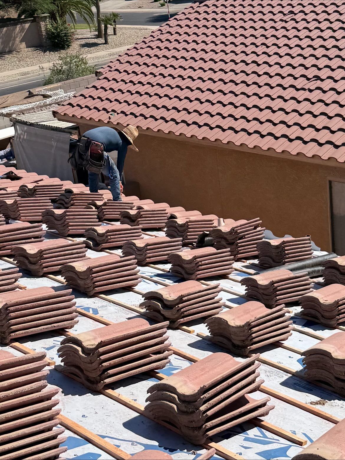 Roofer installing red clay tiles on a rooftop. Tiles are stacked on the roof beside the worker and the completed roof.
