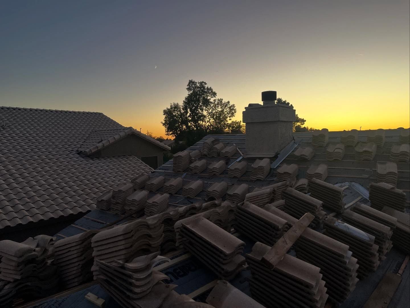 Rooftop with stacked tiles, chimney, tree against sunset sky.