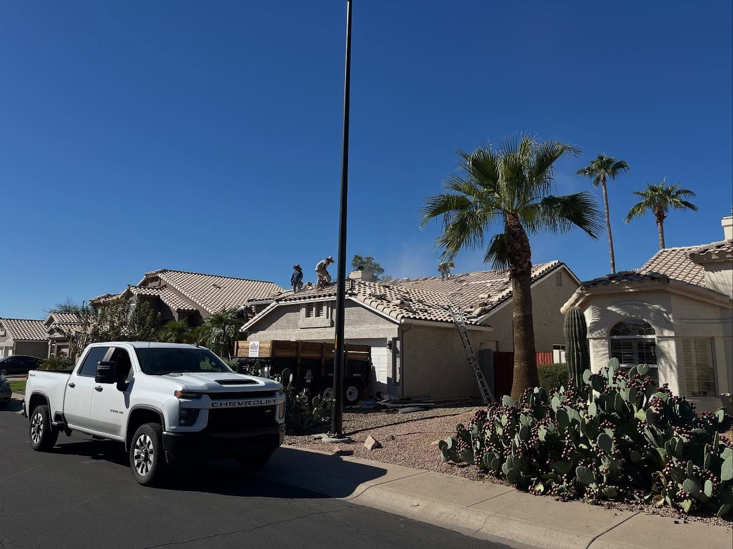 White pickup truck on a sunny residential street with workers on a roof, palm trees, and clear blue sky.