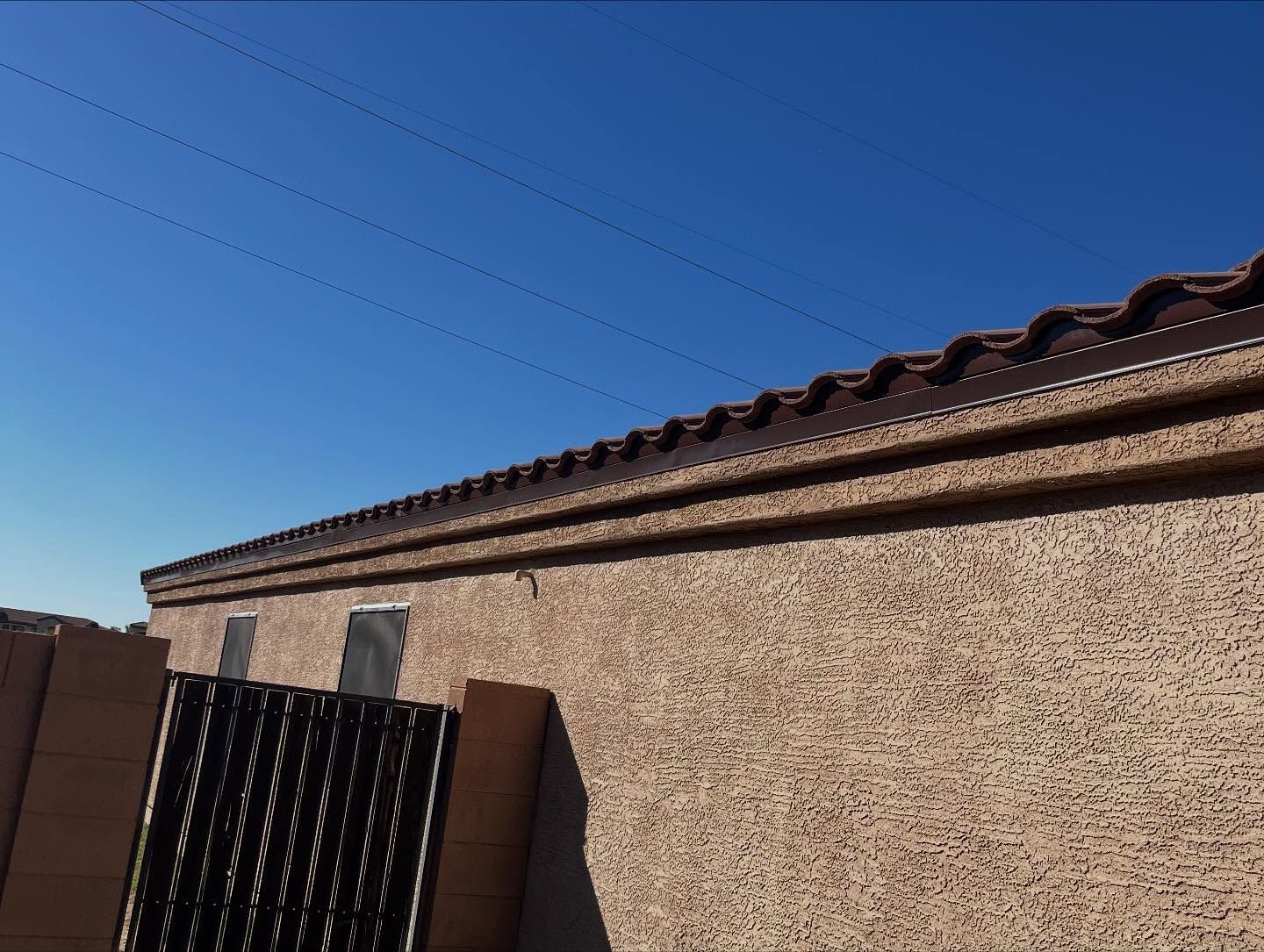 Tan stucco wall with brown trim and tile roof against a bright blue sky.