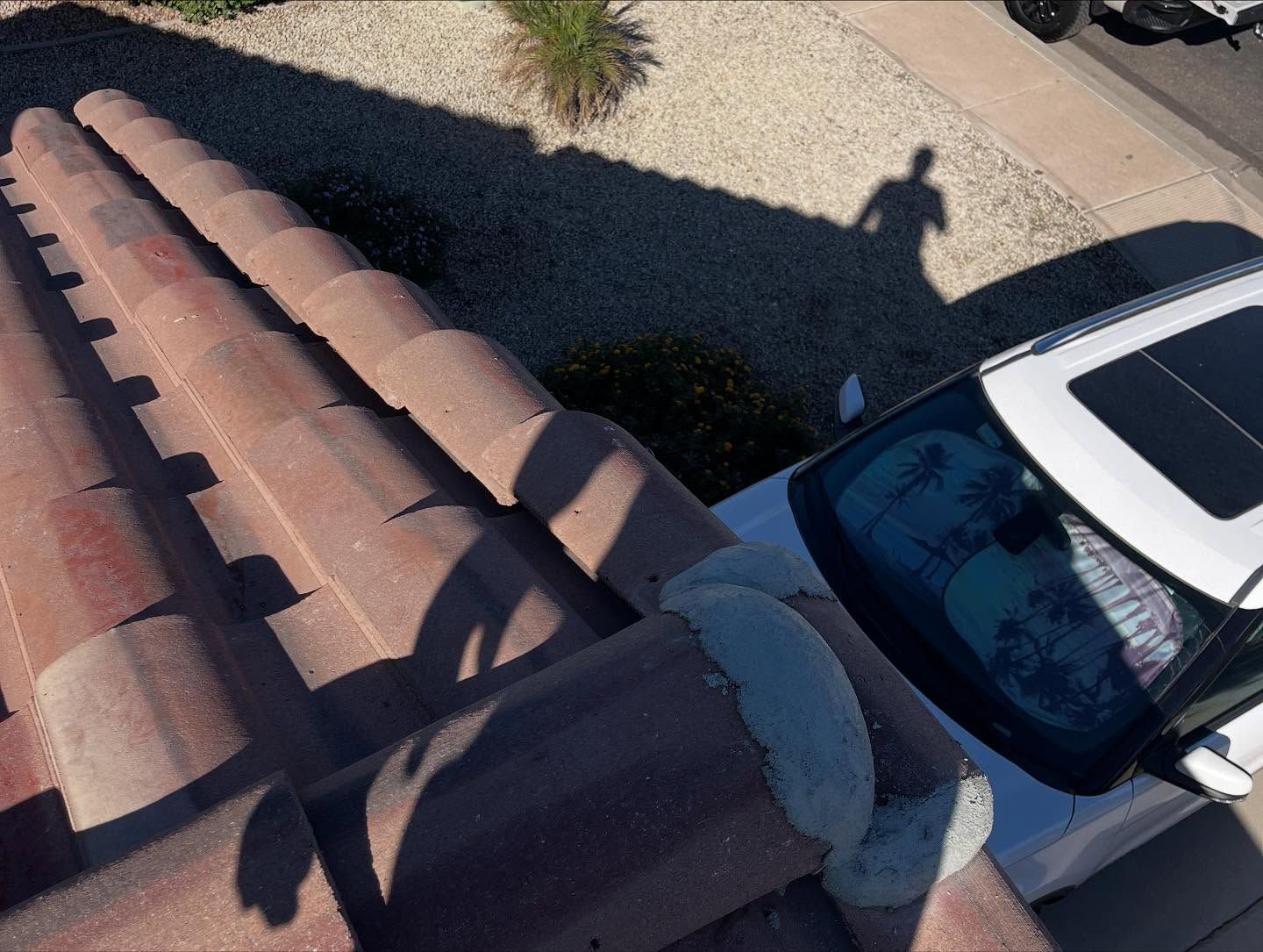 Overhead view of a terracotta tiled roof next to a white car. A shadow of a person is cast on gravel.