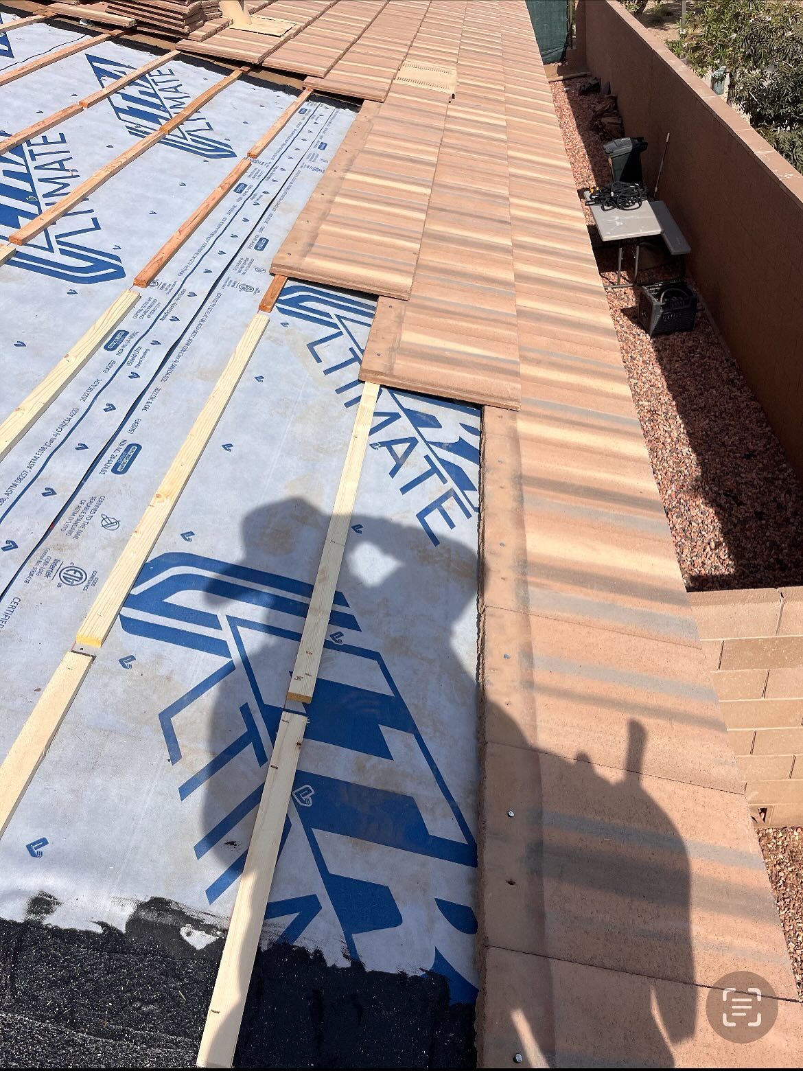 Roofing tiles partially installed on a roof, with underlayment visible, and shadow of a person.