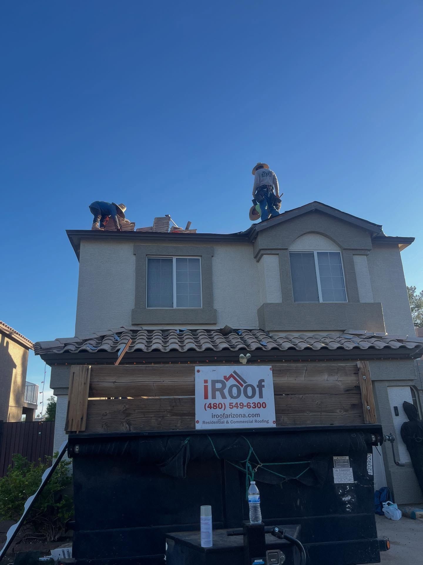 Roofers working on a two-story house with a dumpster in the foreground. Bright blue sky. iRoof sign visible.