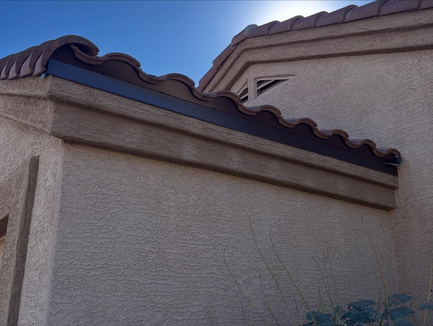 Tan stucco wall with brown roof tiles and trim against a bright sky.