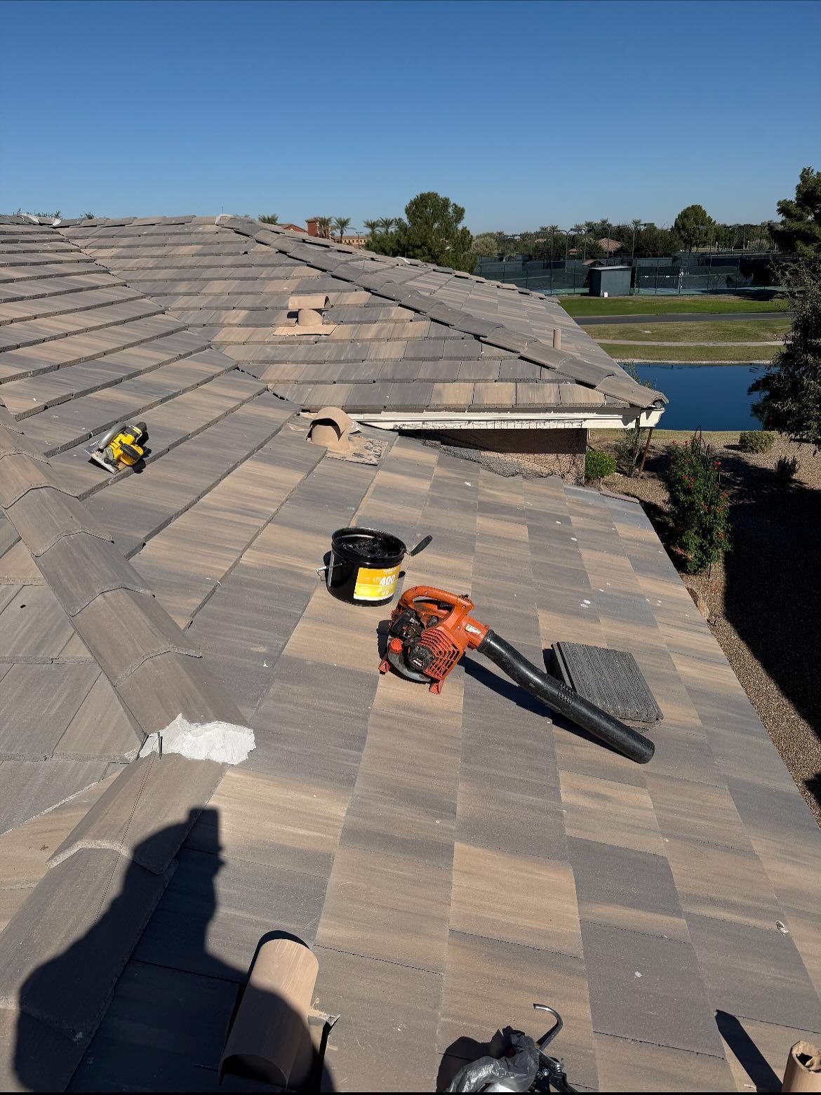 Rooftop with tools: blower, bucket. Brown tile roof, sunny day, clear blue sky. Houses and water in the background.