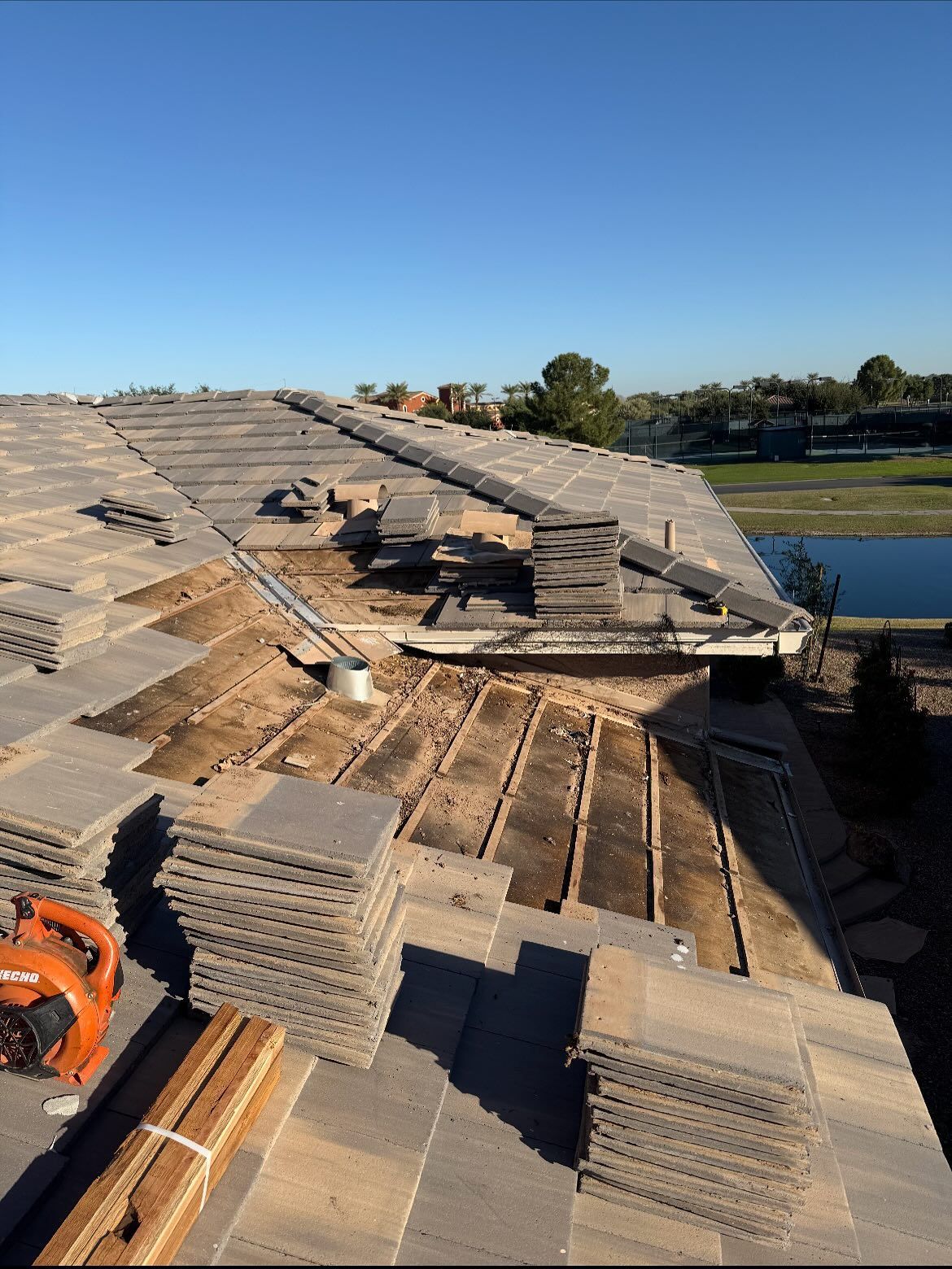 Rooftop under construction; stacks of gray tiles, exposed wooden structure, blue sky, and a lake.
