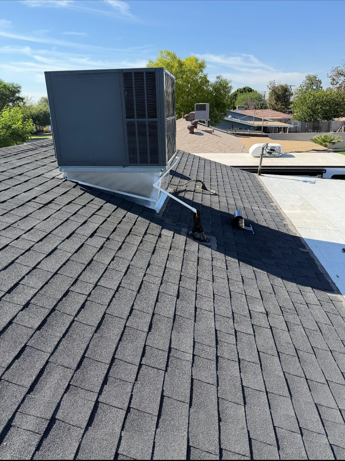 View of a shingled roof with a large gray box on top, against a blue sky with some trees.